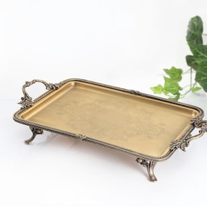 May include: A rectangular gold-toned metal tray with an ornate, dark bronze-colored border and matching handles on each side. The tray features four decorative feet and a subtle, etched floral pattern centered on the flat, smooth surface.