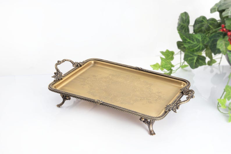 May include: A rectangular gold-toned metal tray with an ornate, dark bronze-colored border and matching handles on each side. The tray features four decorative feet and a subtle, etched floral pattern centered on the flat, smooth surface.