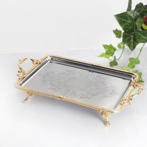 May include: A rectangular silver-toned serving tray with a reflective mirrored surface featuring an intricate etched floral pattern. The tray is framed by an ornate gold-toned metal border with decorative handles and four small claw-style feet.