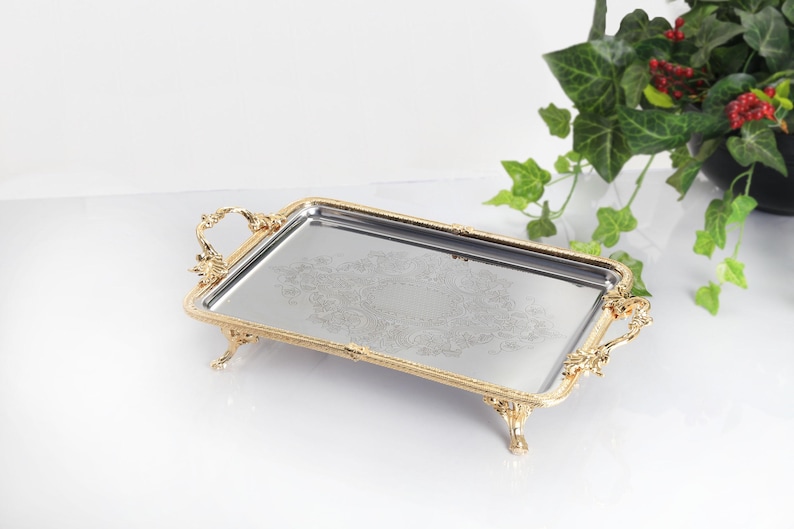 May include: A rectangular silver-toned serving tray with a reflective mirrored surface featuring an intricate etched floral pattern. The tray is framed by an ornate gold-toned metal border with decorative handles and four small claw-style feet.