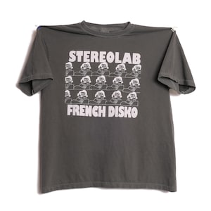 May include: A dark grey t-shirt featuring the words "STEREOLAB" and "FRENCH DISKO" in white, block lettering. Between the text is a grid of white line drawings of a person wearing a hat. The t-shirt is displayed against a white backdrop.