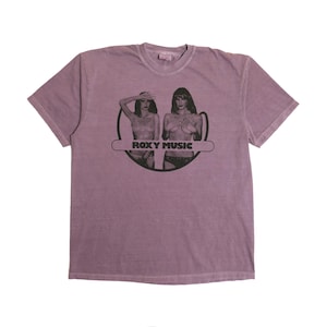 May include: Mauve-colored t-shirt featuring a black and white graphic of two women and the text "ROXY MUSIC" in a horizontal banner. The shirt has short sleeves and a crew neck.