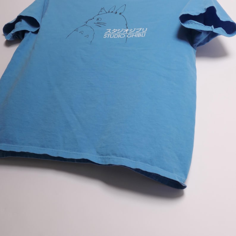 May include: Light blue t-shirt featuring a black outline illustration of a character and Japanese text. The text reads "スタジオジブリ STUDIO GHIBLI". The shirt is made of a soft material.