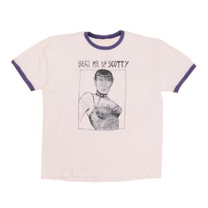 Może przedstawiać: Jasnoróżowy t-shirt typu ringer z ciemnofioletowym wykończeniem przy kołnierzu i rękawach. Na środku znajduje się czarno-biała grafika przedstawiająca postać ze spiczastymi uszami w obroży z ćwiekami i gorsecie, z napisem BEAT ME UP SCOTTY powyżej.