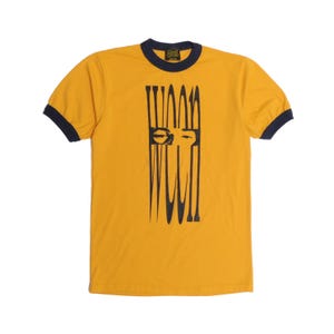 Puede incluir: Camiseta de color mostaza con ribetes azul marino. La camiseta presenta un diseño gráfico negro de una cara con líneas verticales. El texto "WANT" está impreso en el cuello.