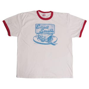 以下が含まれることがあります： 赤色の襟と袖のトリムが付いた白いリンガーTシャツ。前面には、フォークが付いたお皿に乗ったパイの青いグラフィックと、青い長方形のボックスに「Elliott Smith」というテキストが表示されています。