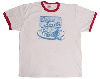 May include: White ringer t-shirt with red trim on the collar and sleeves. The front features a blue graphic of a pie on a plate with a fork, and the text "Elliott Smith" in a blue rectangular box.