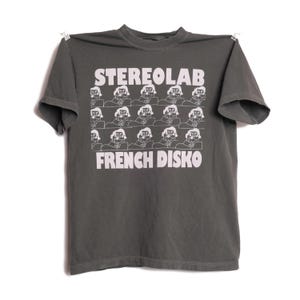 May include: Dark grey t-shirt featuring the text "STEREOLAB" above a grid of illustrated figures and the words "FRENCH DISKO" below. The shirt is suspended against a white backdrop.
