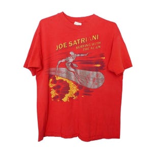 May include: A red short-sleeve t-shirt featuring a graphic of a silver figure surfing on a board. The text JOE SATRIANI SURFING WITH THE ALIEN is printed in yellow and white above the figure, with orange and yellow abstract shapes below.