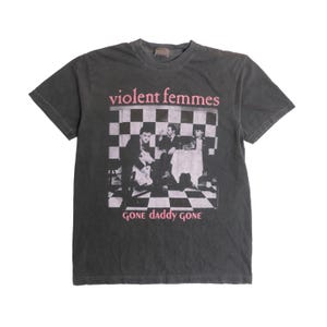 May include: Dark grey t-shirt with the pink text "violent femmes" above a black and white image of the band. The text "GONE daddy GONE" is printed below. The shirt has a crew neck and short sleeves, a classic design.