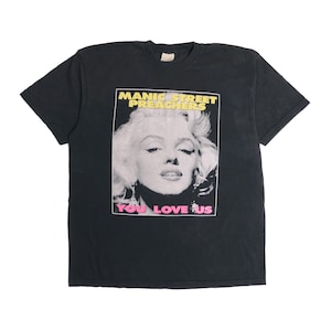 Könnte beinhalten: Schwarzes T-Shirt mit einem Aufdruck von Marilyn Monroe. Der Text "MANIC STREET PREACHERS" befindet sich über dem Bild und "YOU LOVE US" darunter. Das Bild ist gelb und rosa umrandet.