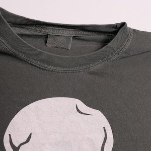 K&ouml;nnte beinhalten: Nahaufnahme eines dunkelgrauen T-Shirts mit Rundhalsausschnitt. Das Shirt hat ein gro&szlig;es, wei&szlig;es Grafikdesign auf der Vorderseite. Der Stoff scheint weich und leicht abgenutzt zu sein.