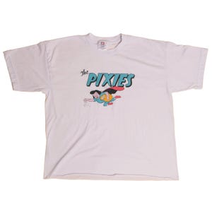 Può includere: T-shirt bianca con il nome della band "The Pixies" in lettere turchesi e nere, e una grafica a cartoni animati di una figura volante. La figura indossa un abito blu, un mantello rosso e ha i capelli neri. La maglietta è a maniche corte.