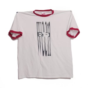 Puede incluir: Camiseta blanca con cuello y ribetes de manga rojos. La camiseta presenta un diseño gráfico negro de una cara con líneas verticales. El texto "FAR WEST" está impreso en el cuello.