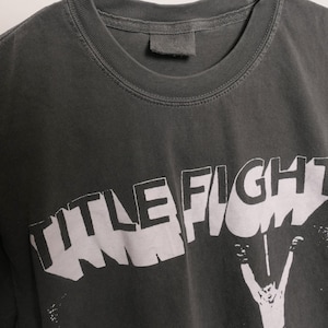 May include: A dark gray t-shirt with a white graphic print of the word "TITLE FIGHT" in a bold, stylized font. The graphic is partially obscured by a figure with raised arms.