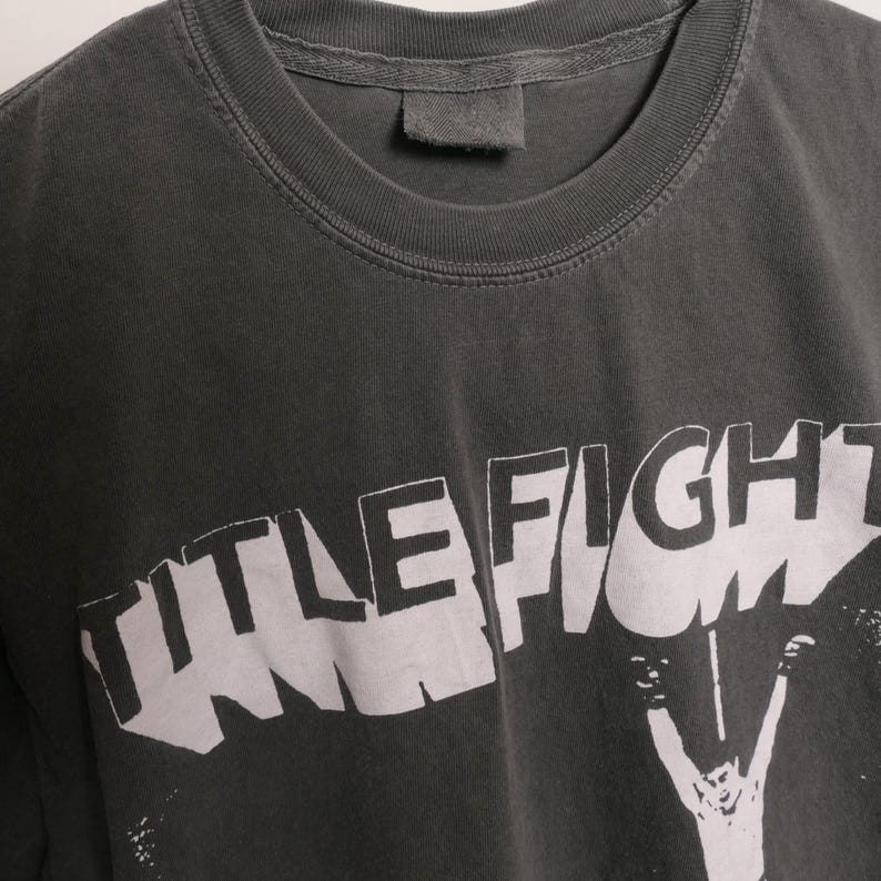 May include: A dark gray t-shirt with a white graphic print of the word "TITLE FIGHT" in a bold, stylized font. The graphic is partially obscured by a figure with raised arms.