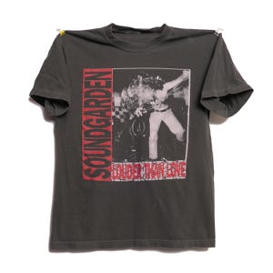 以下が含まれることがあります： 左側に赤い文字でSOUNDGARDEN、下部に赤い文字でLOUDER THAN LOVEとプリントされた、チャコールグレーの半袖Tシャツ。中央には、パフォーマンス中の人物を写した白黒写真のグラフィックがデザインされています。