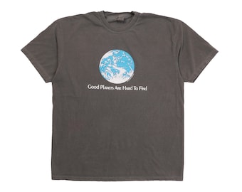 May include: A charcoal gray short-sleeve t-shirt featuring a circular graphic of the Earth in shades of blue and white. Below the graphic, the text reads "Good Planets Are Hard To Find" in a white, stylized serif font centered on the chest.