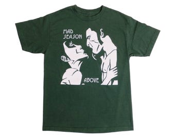 Könnte beinhalten: Dunkelgrünes T-Shirt mit einem weißen Grafikdesign, das zwei stilisierte Figuren zeigt, die sich gegenüberstehen. Der Text "MAD SEASON" steht über den Figuren und "ABOVE" darunter. Das T-Shirt ist aus weichem Material.