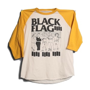 Puede incluir: Camiseta de béisbol color crema con mangas amarillas. La camiseta presenta el texto "BLACK FLAG" sobre un gráfico de varias figuras. Debajo de las figuras hay líneas verticales negras. La camiseta está hecha de un material suave.