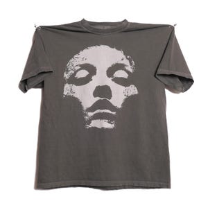Könnte beinhalten: Ein graues T-Shirt mit einer weißen Grafik eines Gesichts mit geschlossenen Augen und einer markanten Nase und Mund. Das T-Shirt besteht aus einem weichen Material und hat kurze Ärmel und einen Rundhalsausschnitt.