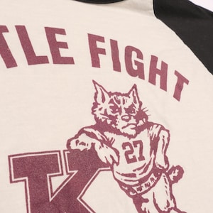 May include: Cream-colored baseball style shirt with black sleeves. The shirt features the text "LITTLE FIGHT" above a maroon illustration of a wildcat wearing a jersey with the number 27. The letter "K" is also visible.
