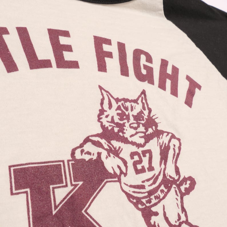 May include: Cream-colored baseball style shirt with black sleeves. The shirt features the text "LITTLE FIGHT" above a maroon illustration of a wildcat wearing a jersey with the number 27. The letter "K" is also visible.
