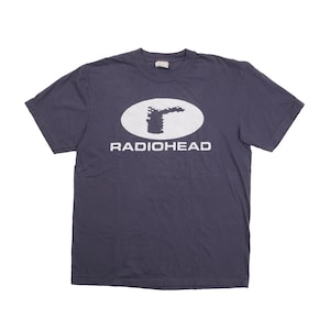 Puede incluir: Una camiseta de manga corta de color gris oscuro con un gráfico ovalado blanco en el pecho. Dentro del óvalo hay una forma oscura, dentada y abstracta sobre la palabra RADIOHEAD impresa en una fuente blanca, negrita y sin remates.