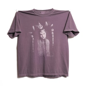 May include: A purple t-shirt with a faded image of a woman with a crown on her head. The image is in shades of gray and white.