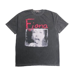 May include: A charcoal gray short-sleeve t-shirt featuring a black and white photographic print of a person with their mouth open and tongue out. The text Fiona Apple is printed in a distressed red font above the central graphic image.