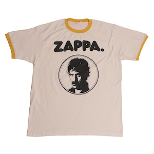 Puede incluir: Camiseta color crema con cuello y ribetes de manga amarillos. La palabra "ZAPPA." está impresa en negro sobre un retrato circular de un hombre con bigote. Una camiseta gráfica de estilo vintage.