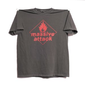 May include: Dark gray t-shirt featuring a red diamond with a flame graphic and the words "massive attack" in red. The shirt is made of a soft material and has short sleeves.