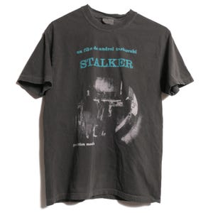 May include: A dark gray t-shirt with a faded image of a scene from the film "Stalker" by Andrei Tarkovsky. The text "un film de andrei tarkovsky" and "STALKER" are printed in blue on the shirt.