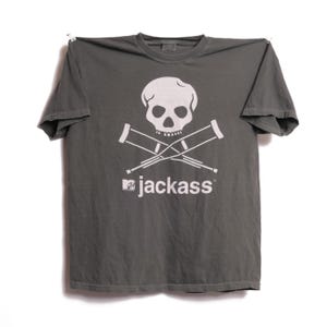 May include: A gray t-shirt featuring a white skull and crossbones design with crossed crutches. Below the graphic is the text "jackass" with the MTV logo. The shirt is hanging against a white background.
