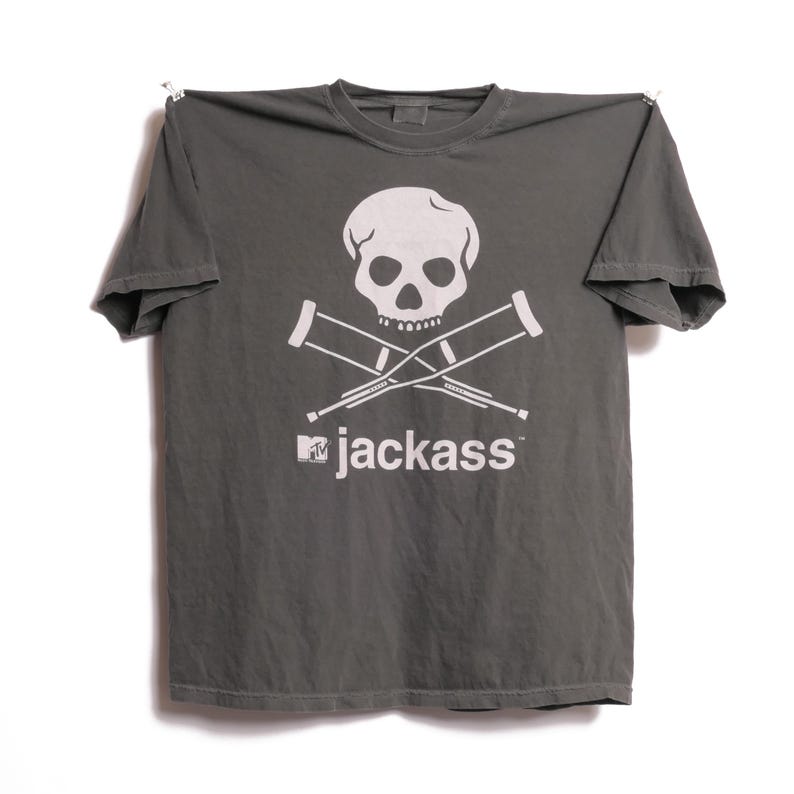 K&ouml;nnte beinhalten: Ein graues T-Shirt mit einem wei&szlig;en Totenkopf- und gekreuzten Knochen-Design mit gekreuzten Kr&uuml;cken. Der Text "jackass" mit dem MTV-Logo ist darunter aufgedruckt. Das T-Shirt h&auml;ngt vor einem wei&szlig;en Hintergrund.