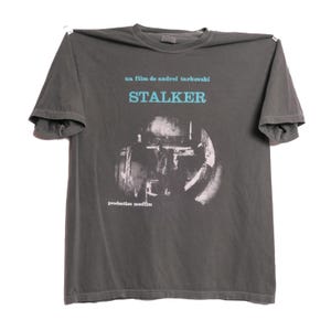 May include: A dark gray t-shirt with a white graphic print of a scene from the film "Stalker" by Andrei Tarkovsky. The text "un film de Andrei Tarkovsky" and "STALKER" are printed in blue above the image. The text "production mosfilm" is printed in white below the image.