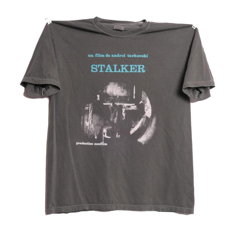 May include: A dark gray t-shirt with a white graphic print of a scene from the film "Stalker" by Andrei Tarkovsky. The text "un film de Andrei Tarkovsky" and "STALKER" are printed in blue above the image. The text "production mosfilm" is printed in white below the image.