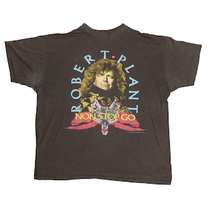 May include: A dark gray t-shirt featuring a portrait of Robert Plant with the text "ROBERT PLANT" in blue and "NON STOP GO" in yellow. The graphic includes red and yellow accents.