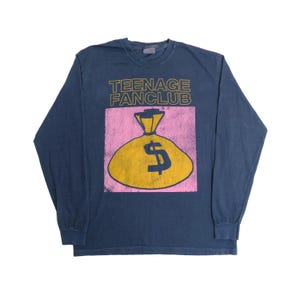 Puede incluir: Camiseta de manga larga azul marino con el texto "TEENAGE FANCLUB" sobre un gráfico de bolsa de dinero amarilla con un signo de dólar sobre un fondo rosa. La camiseta tiene cuello redondo.