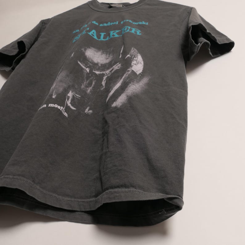 May include: A dark gray t-shirt with a faded image of a person wearing a hat and the text "Andrei Tarkovsky Stalker a most film".