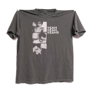 May include: A dark gray t-shirt with a band graphic featuring black and white photos of four people. The band name "YEAH YEAH YEAS" is printed in white letters.