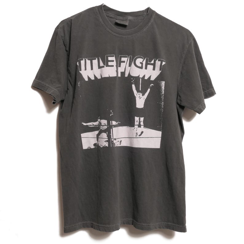 May include: A dark gray t-shirt with a white graphic print of two wrestlers in a ring. The text "TITLE FIGHT" is printed above the wrestlers.