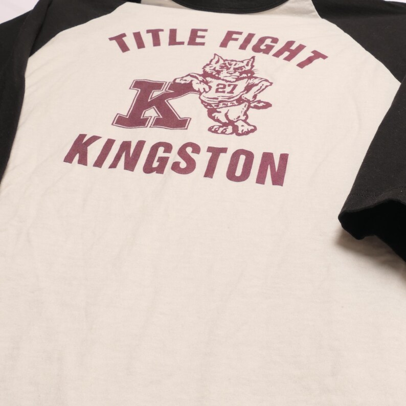May include: Cream-colored baseball style t-shirt with black sleeves. The shirt features the text "TITLE FIGHT" above a maroon graphic of a cat wearing a jersey, and the word "KINGSTON" below.