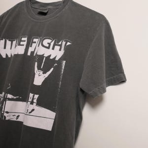 May include: A dark gray t-shirt with a white graphic print of a person in a boxing ring with the text "The Fight" above them.