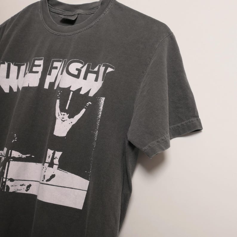 May include: A dark gray t-shirt with a white graphic print of a person in a boxing ring with the text "The Fight" above them.