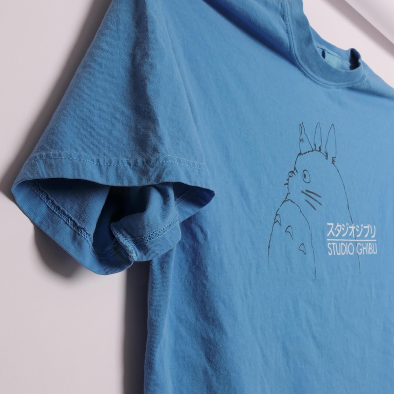 May include: A light blue t-shirt featuring a black graphic of a character from Studio Ghibli, with Japanese text and the English text "STUDIO GHIBLI". The shirt has short sleeves and a crew neck.