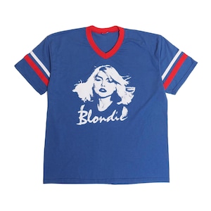 May include: Blue t-shirt with a white graphic of a woman's face and the word "Blondie" in white script. The shirt has a red V-neck collar and red and white stripes on the sleeves.