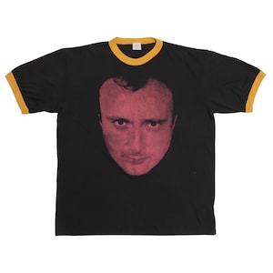 Peut inclure: T-shirt noir avec un graphisme de visage rouge et rose. Le t-shirt a un col et des bordures de manches jaunes. Le graphisme est un gros plan du visage d'une personne.