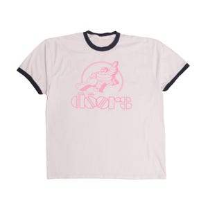 Puede incluir: Camiseta blanca con un gráfico rosa de una figura y la palabra "DOORS". La camiseta tiene cuello y ribetes de manga azul marino. El gráfico está en un círculo.