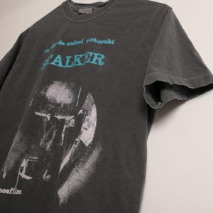 May include: A dark gray t-shirt with a black and white image of a person holding a gun. The text "film by Andrei Tarkovsky STALKER" is printed in blue on the shirt.
